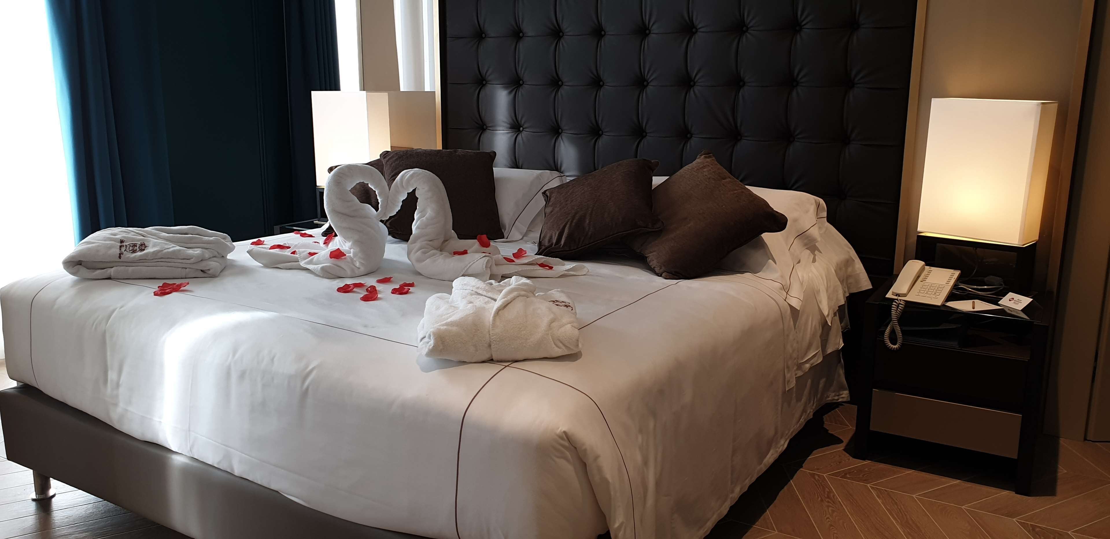 A neatly made bed with towels, pillows, and rose petals. There is a bedside table with a lamp and a phone.