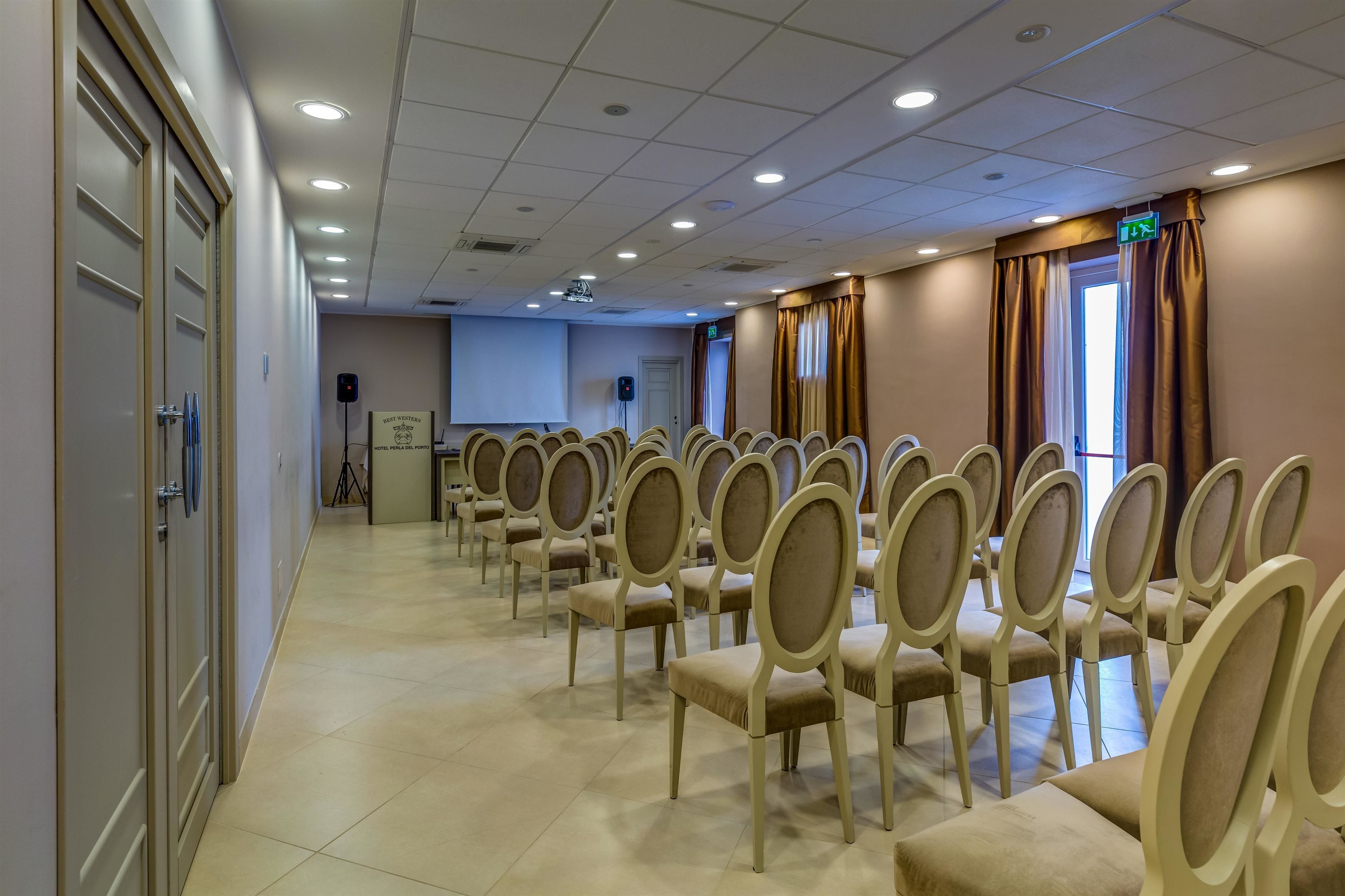 Uma sala de conferências com fileiras de cadeiras bege, uma tela de projetor e um orador.