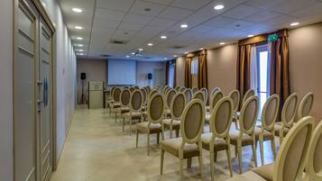 Uma sala de conferências com fileiras de cadeiras bege, uma tela de projetor e um orador.
