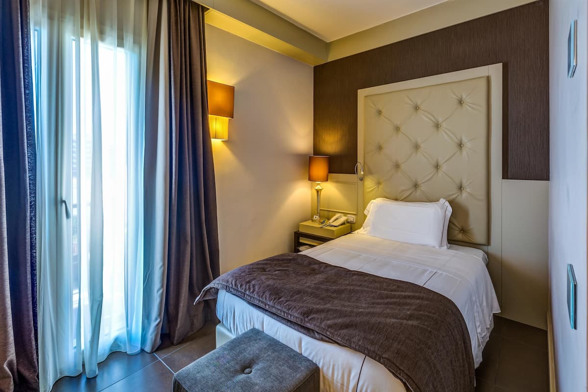 A hotel room with a large bed, a bedside table with a lamp, a telephone, and a window with curtains.