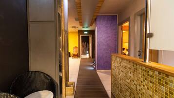 A hotel corridor with a wooden floor, a purple wall, and a tiled wall with a mosaic pattern. There is a wicker chair and a wooden bench along the corridor.