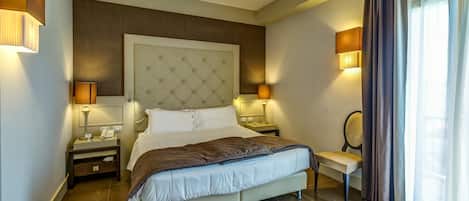 A modern hotel bedroom with a large bed, bedside lamps, a chair, and a view of the outdoors through curtains.