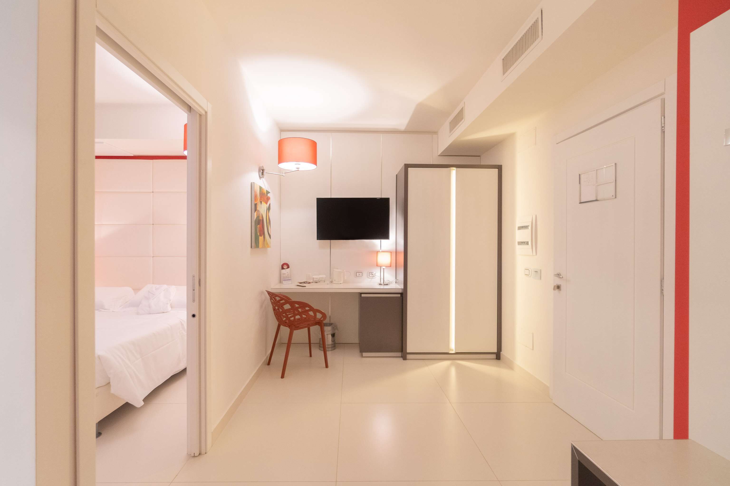Quarto de hotel com cama, escrivaninha, cadeira, televisão e porta.