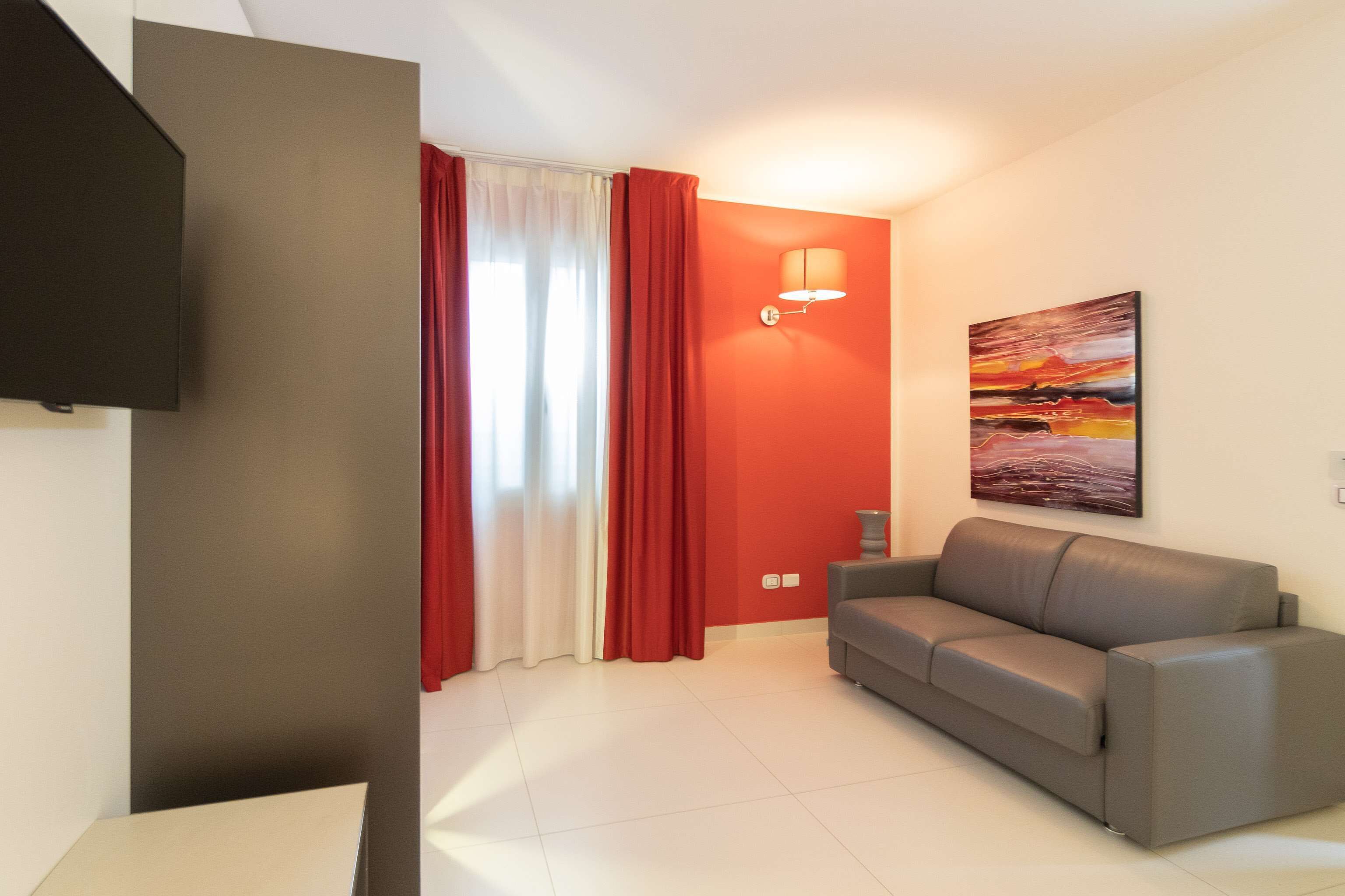 A living room with a grey sofa, red curtains, a wall-mounted lamp, and a painting.