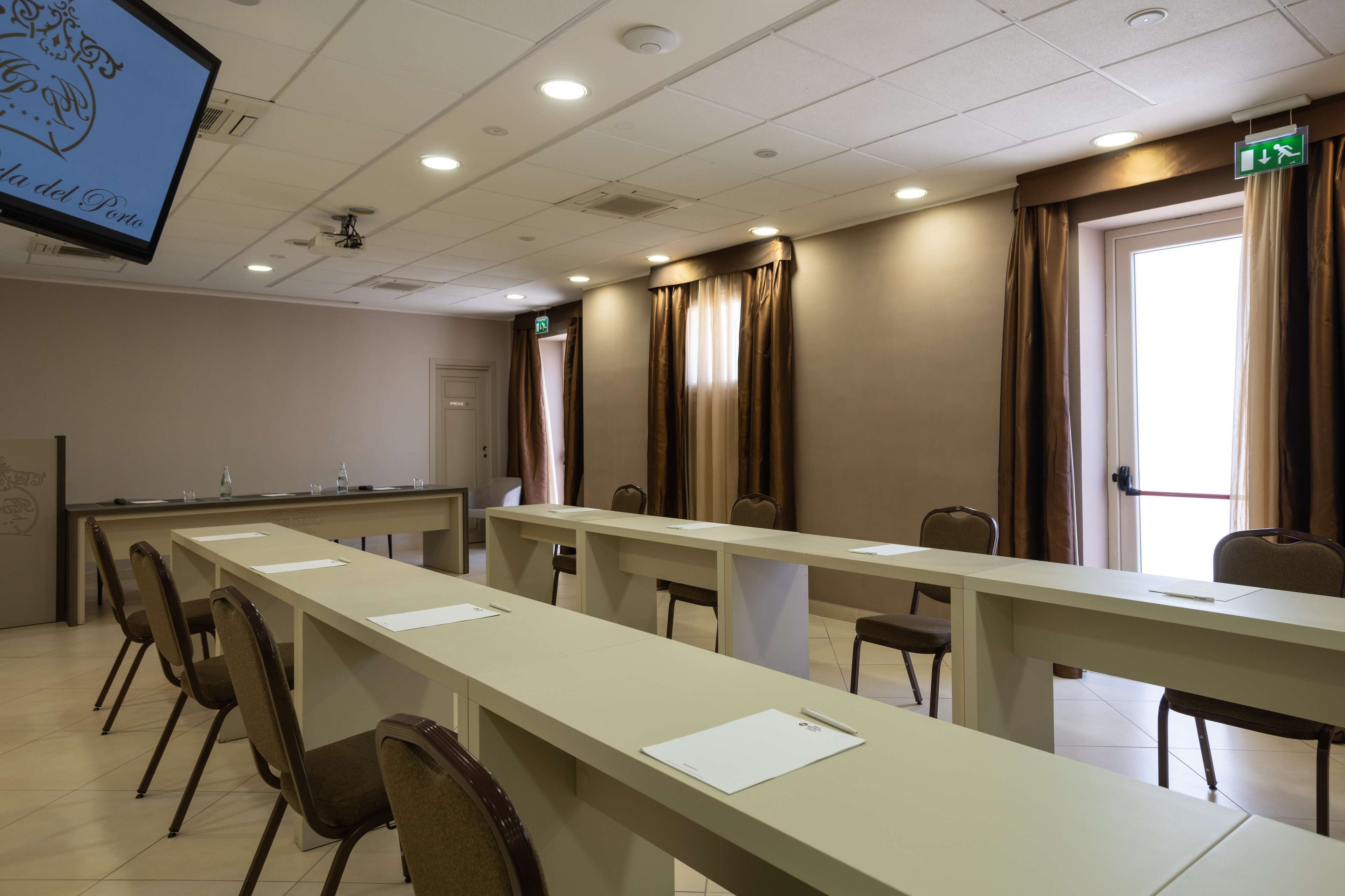 A conference room with long tables and chairs, a projector screen, and an exit sign.