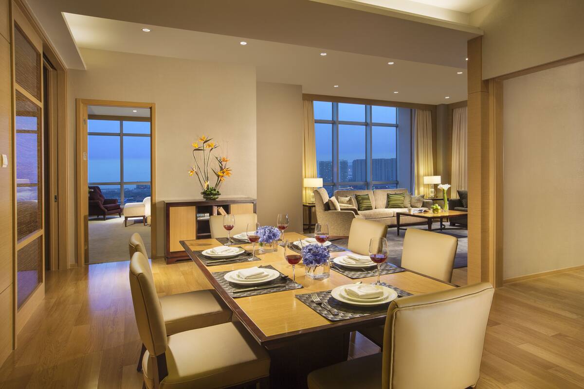 A modern hotel dining area with a large table set for six, a sofa, and a view of the cityscape through large windows.