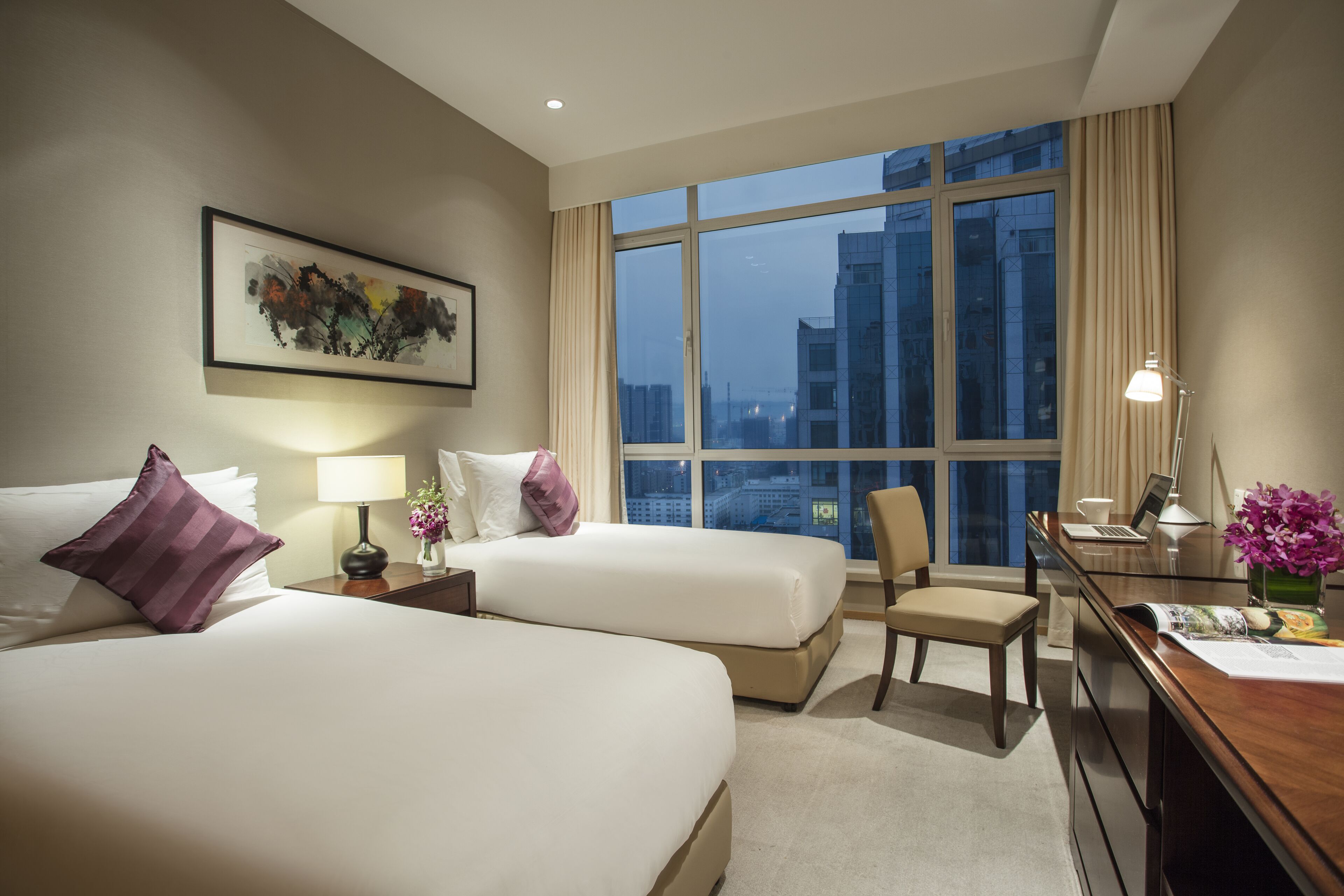 A hotel room with two beds, a desk, a chair, a large window with a city view, and a framed artwork on the wall.