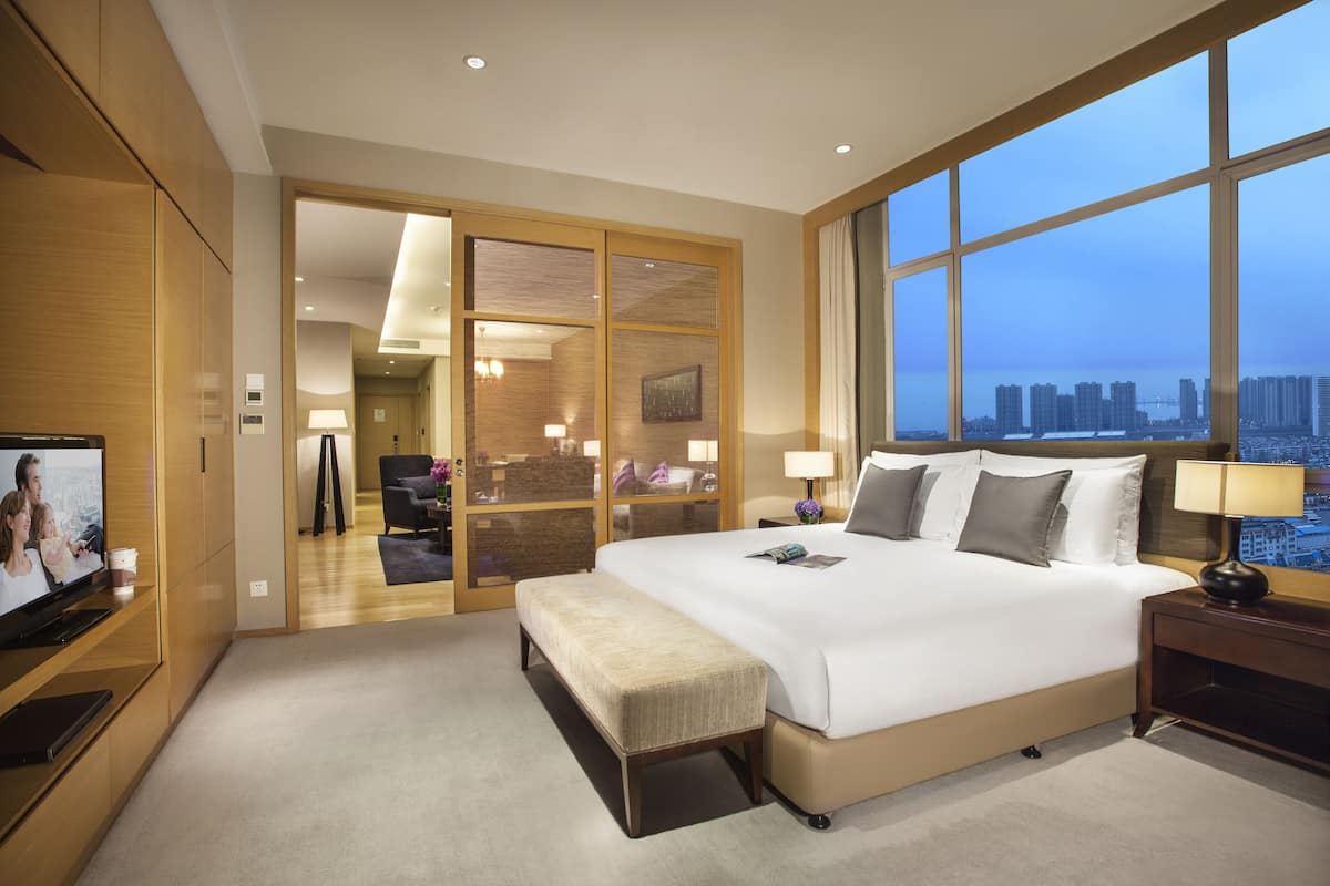 A modern hotel room with a large bed, a TV, a bench, and a view of the city skyline.