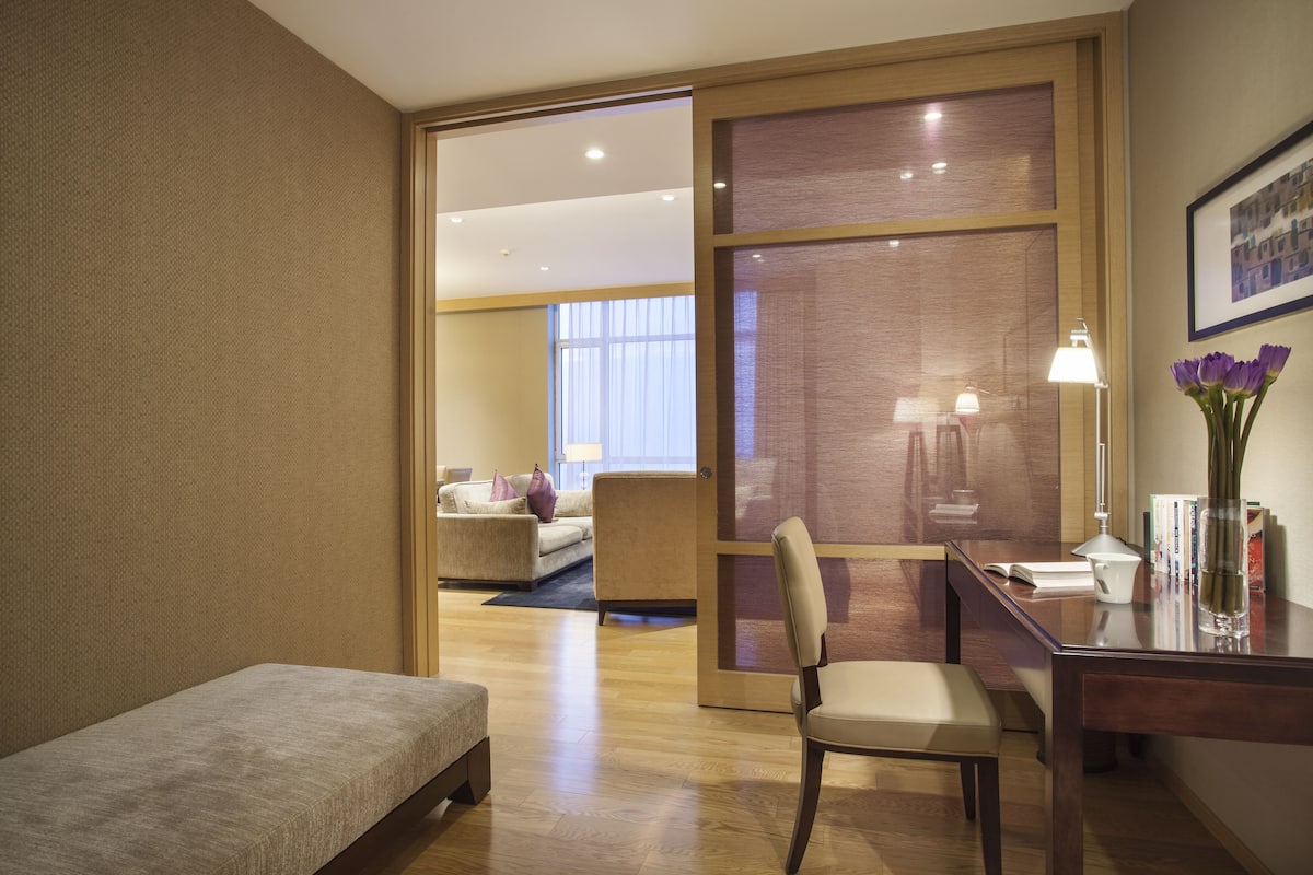 A hotel room with a bed, a desk with a lamp, a chair, and a view of another room with a sofa and a window.
