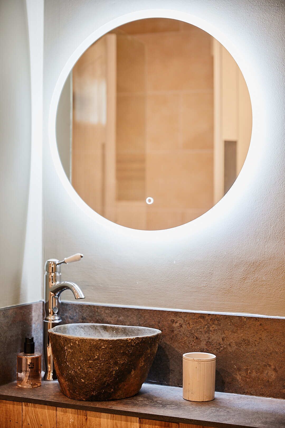A bathroom with a round mirror, a stone sink, and a wooden cup on the counter.
