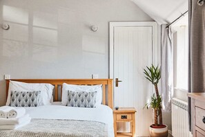 Un lit bien fait, avec du linge de lit blanc et des coussins à motifs, une table de chevet en bois avec un tiroir, une plante en pot et une porte blanche avec une fenêtre sur le côté droit.