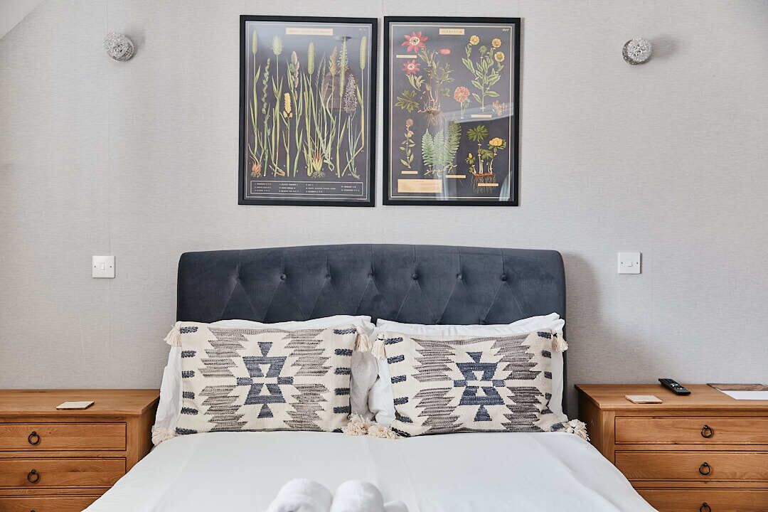 A bedroom with a tufted headboard, two framed botanical prints, patterned pillows, a wooden nightstand, and a bedside table with a remote control.