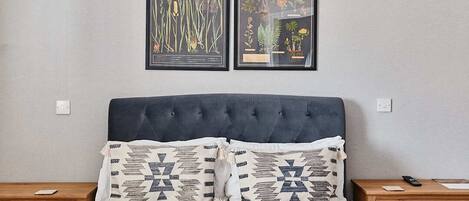 A bedroom with a tufted headboard, two framed botanical prints, patterned pillows, a wooden nightstand, and a bedside table with a remote control.