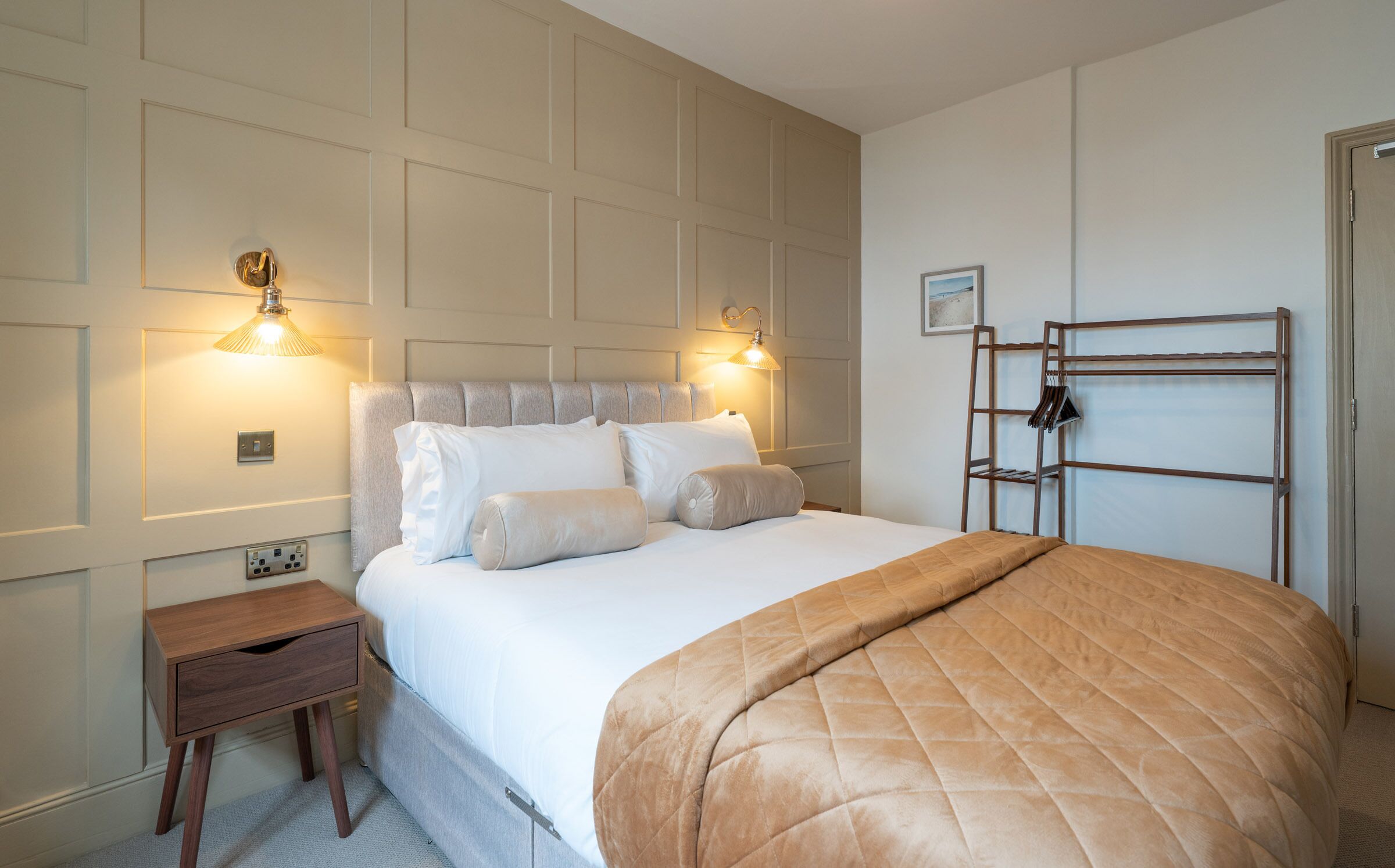Une chambre d’hôtel bien aménagée, avec un grand lit, des lampes de chevet, une petite table de nuit en bois et une étagère en métal.