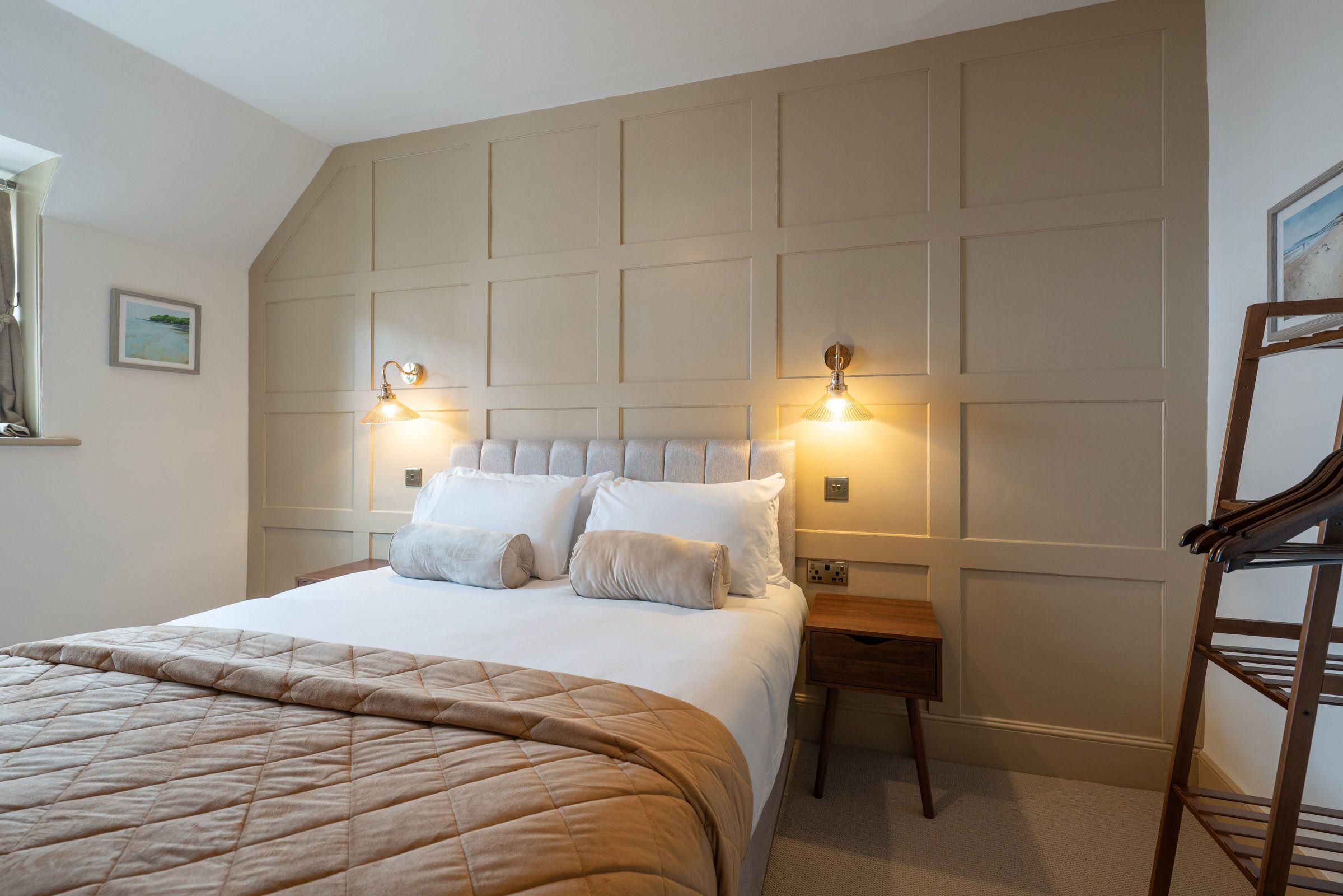 Ein ordentlich bezogenes Bett mit einer beigen Steude, ein Nachttisch aus Holz mit einer Lampe und eine Holzleiter an der Wand.
