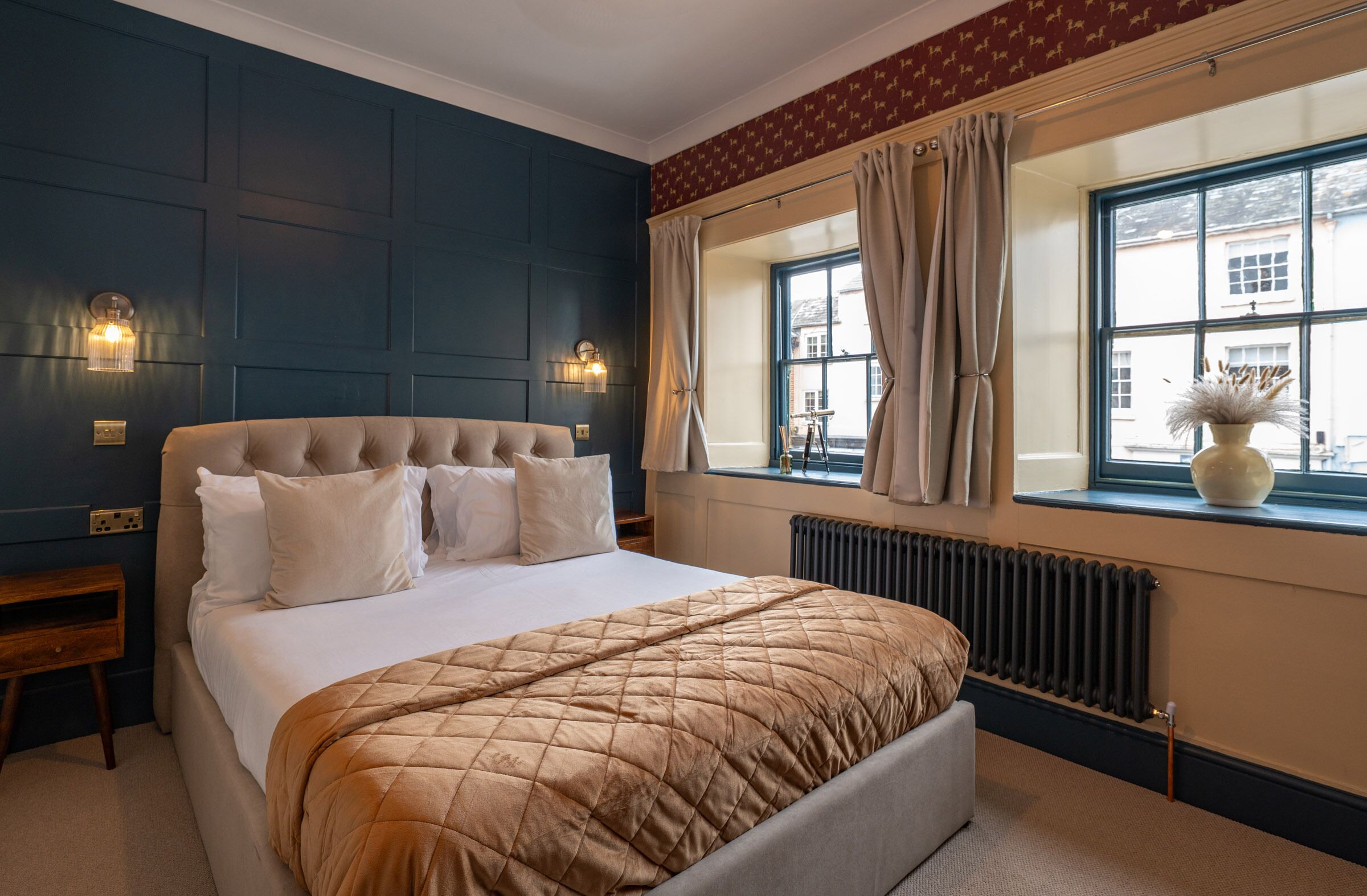 A bedroom with a large bed, two bedside lamps, a window with curtains, and a radiator.
