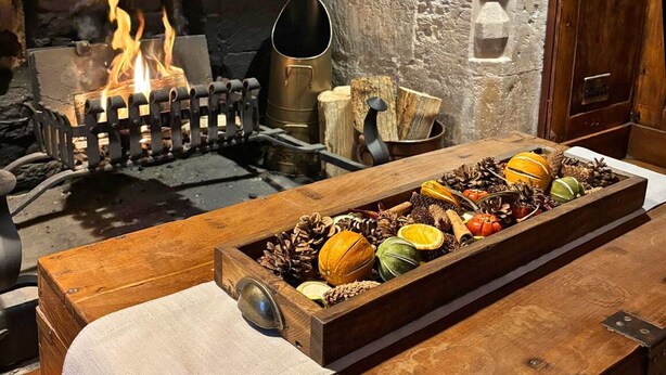 A rustic kitchen with a wood-burning stove, a wooden table with a tray of seasonal fruits and spices, and a stone wall. - The Bugle Coaching Inn (Yarmouth)