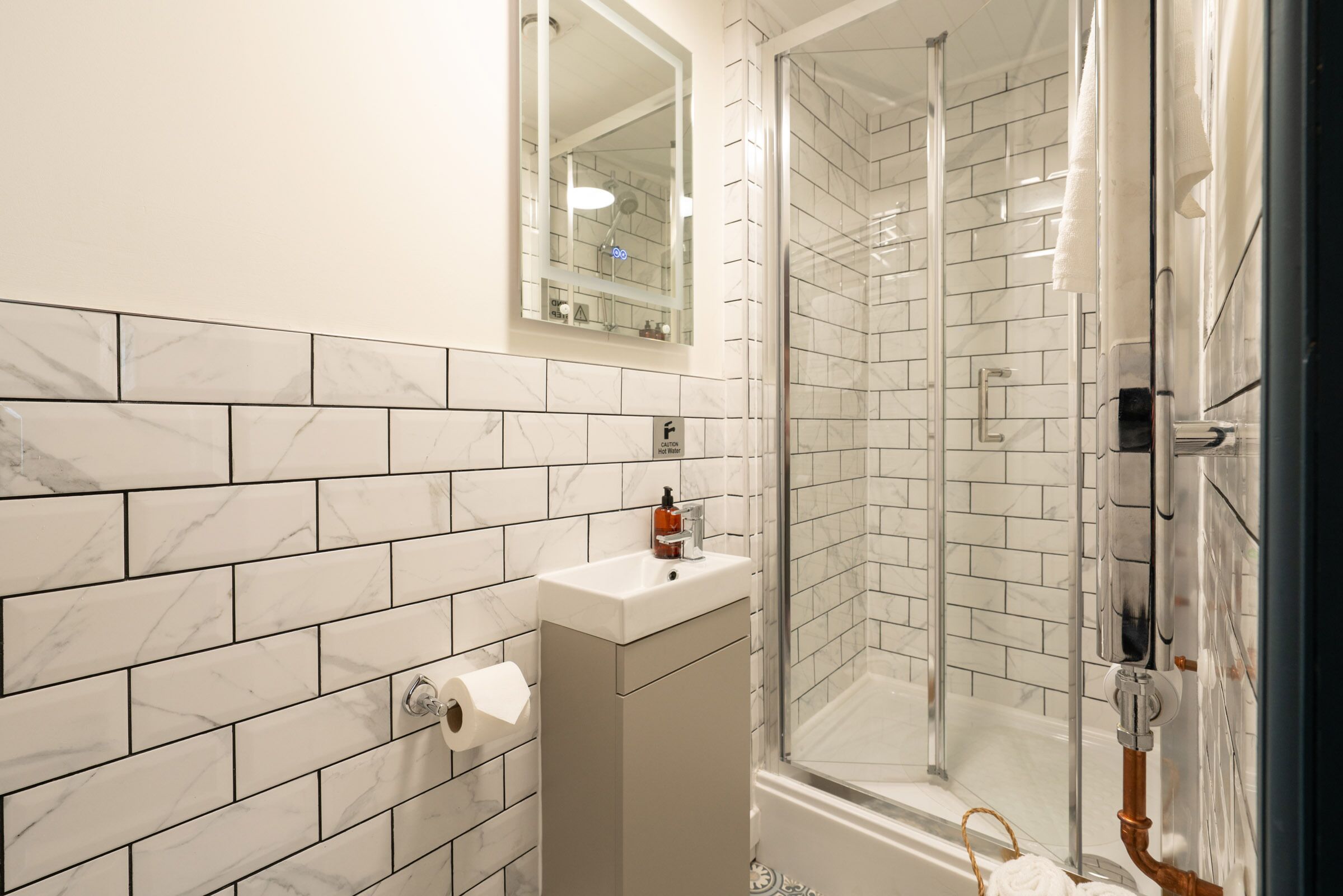 Un bany modern amb rajoles de marbre blanc, una cabina de dutxa de vidre, un lavabo amb dosificador de sabó i un suport per a tovalloles.