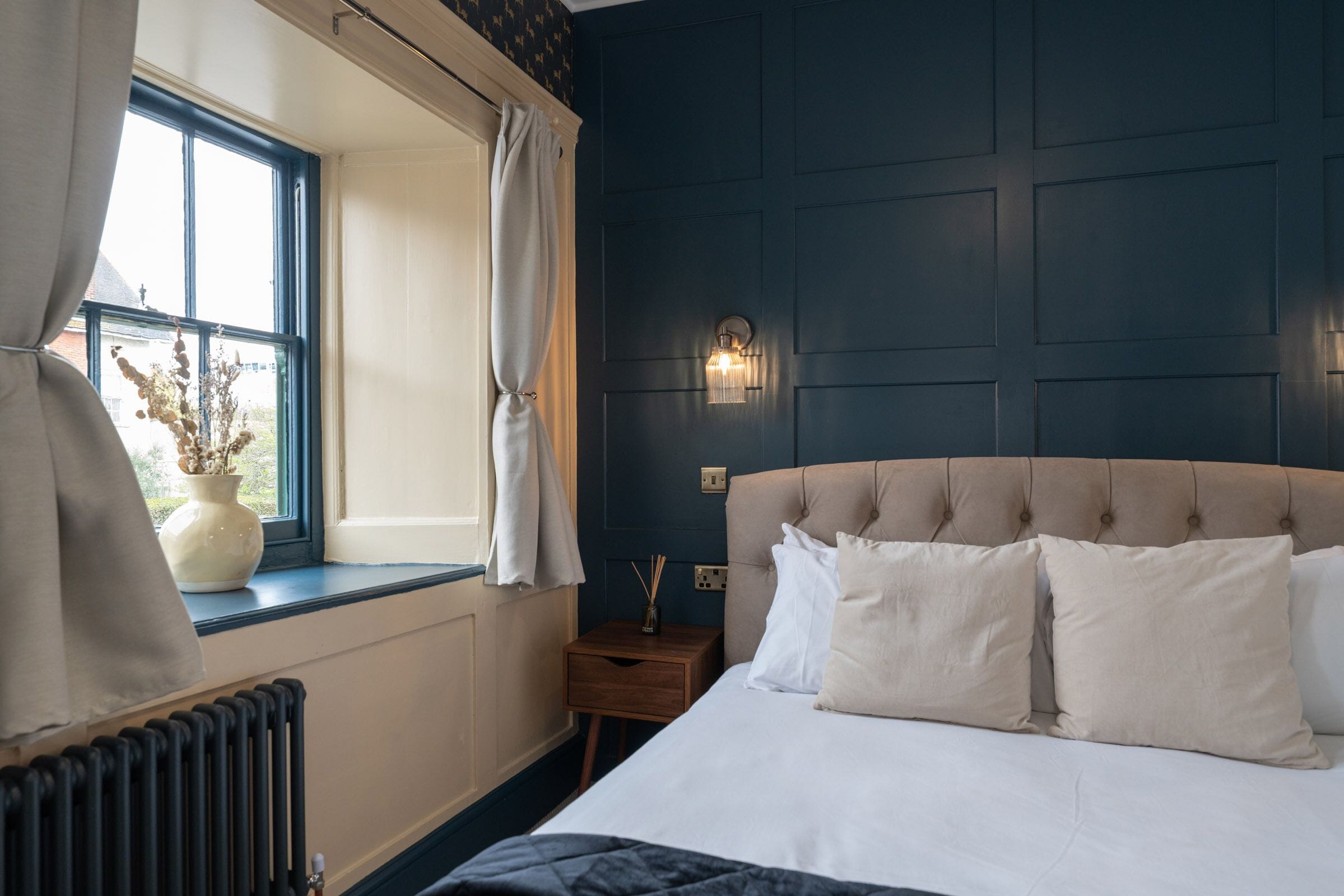 Un dormitorio con una cama grande, una mesita de noche, un ventanal con cortinas y una lámpara de pared.
