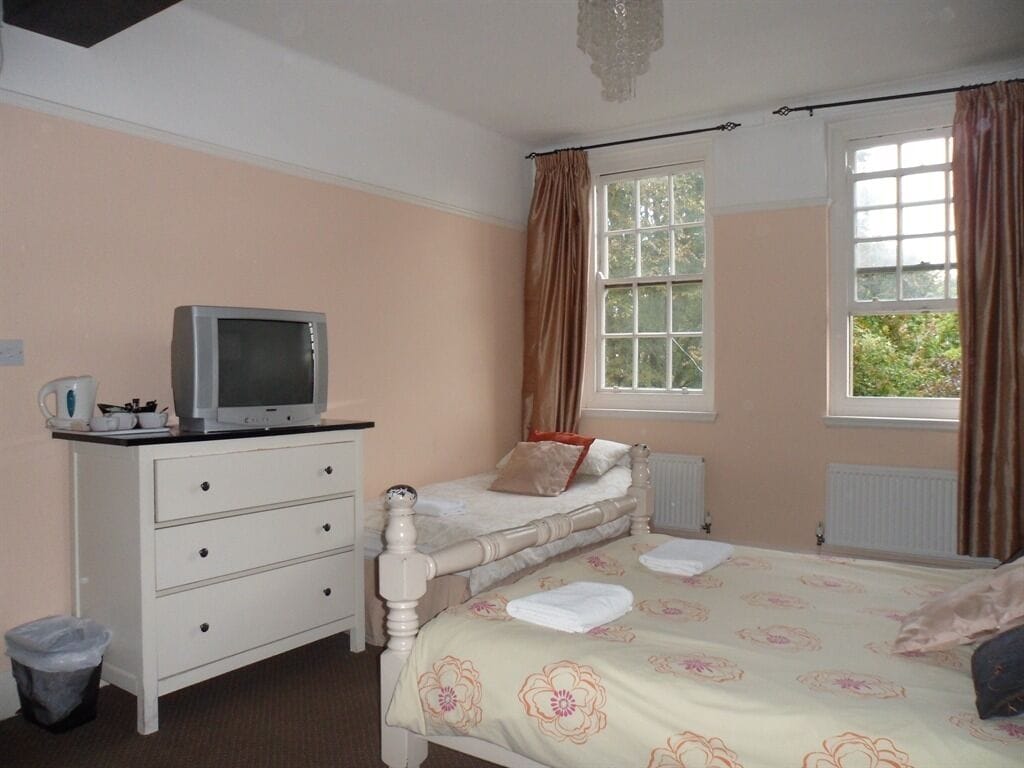Une chambre à coucher comprenant un lit, une commode, un téléviseur, une fenêtre avec des rideaux et un radiateur.