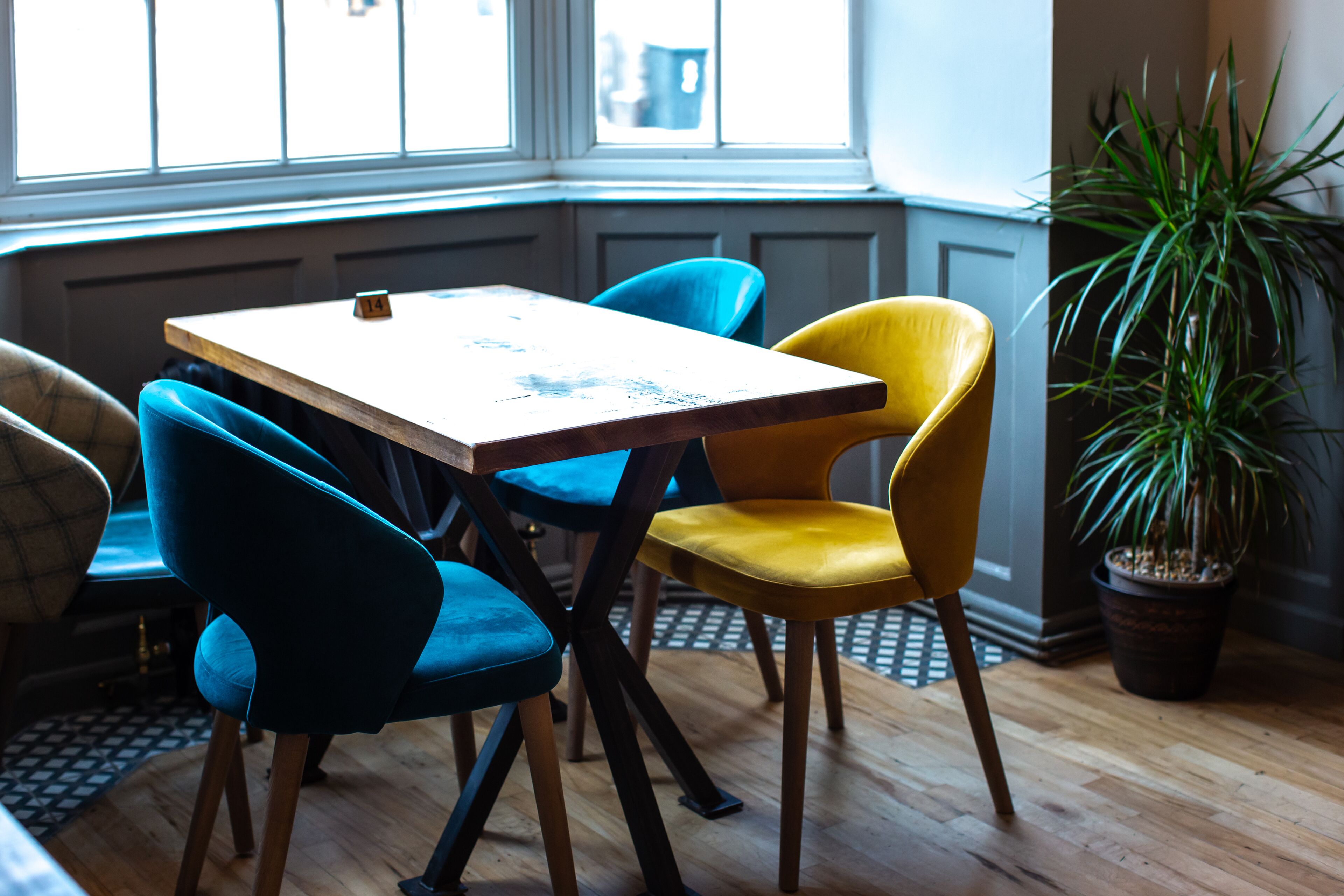A corner seating area with a wooden table and four chairs, one yellow and three blue, in a room with a large window and a potted plant.