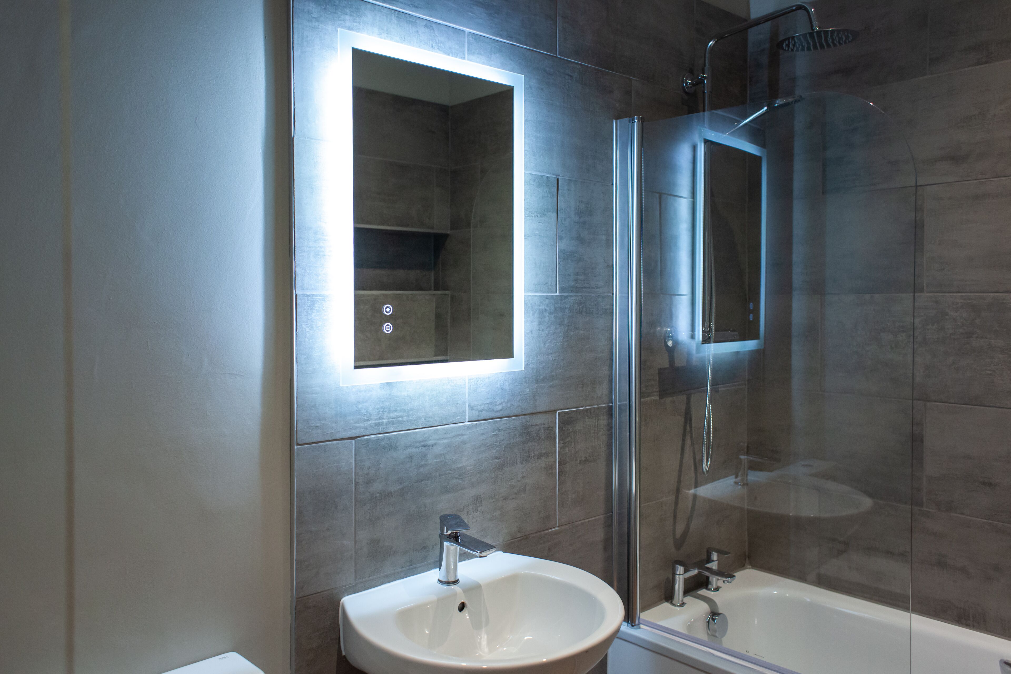 Een moderne badkamer met een glazen douchecabine, een witte wastafel en een spiegel met ingebouwde verlichting.