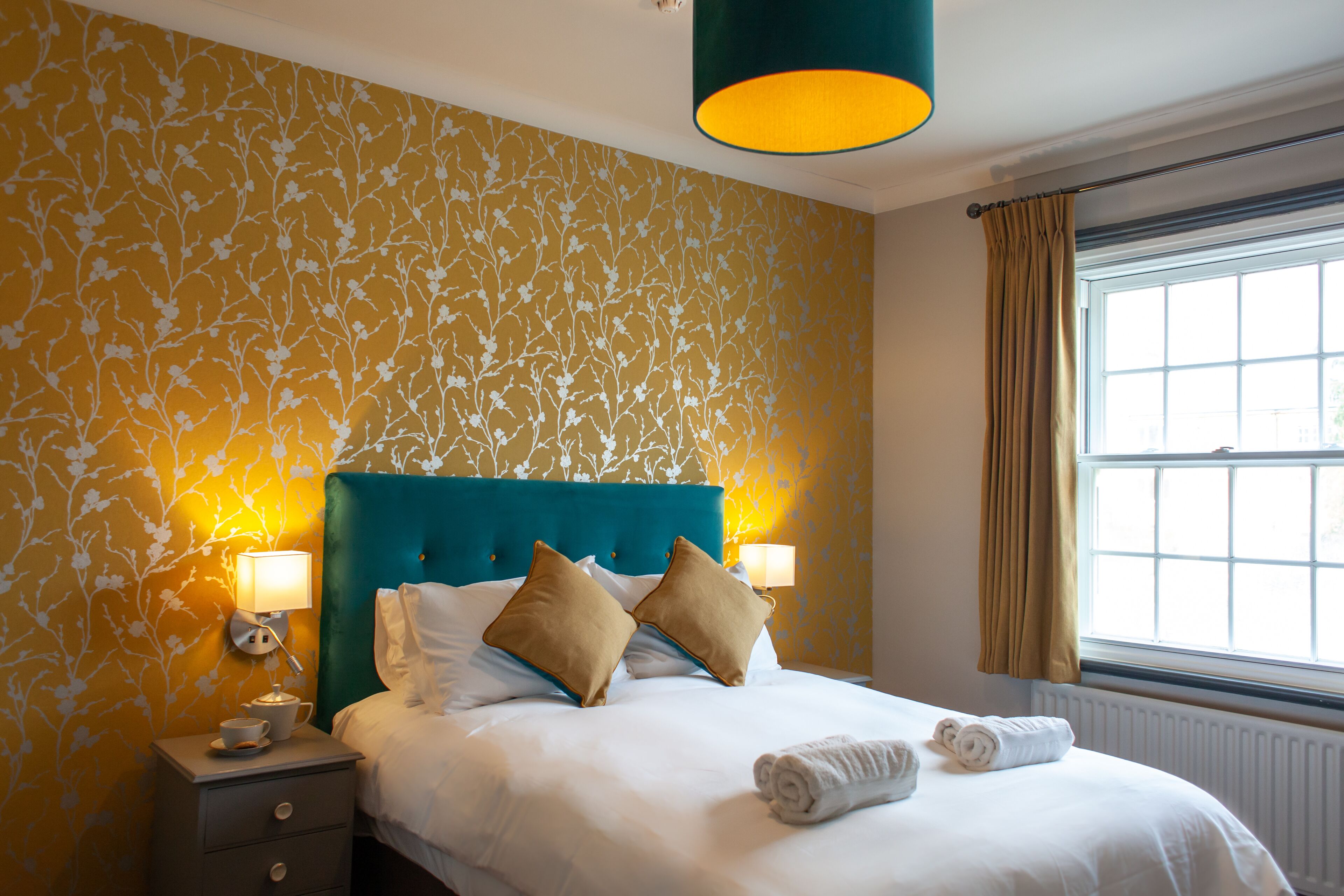 A neatly made bed with white linens and two beige pillows, a bedside table with a lamp, and a window with curtains.