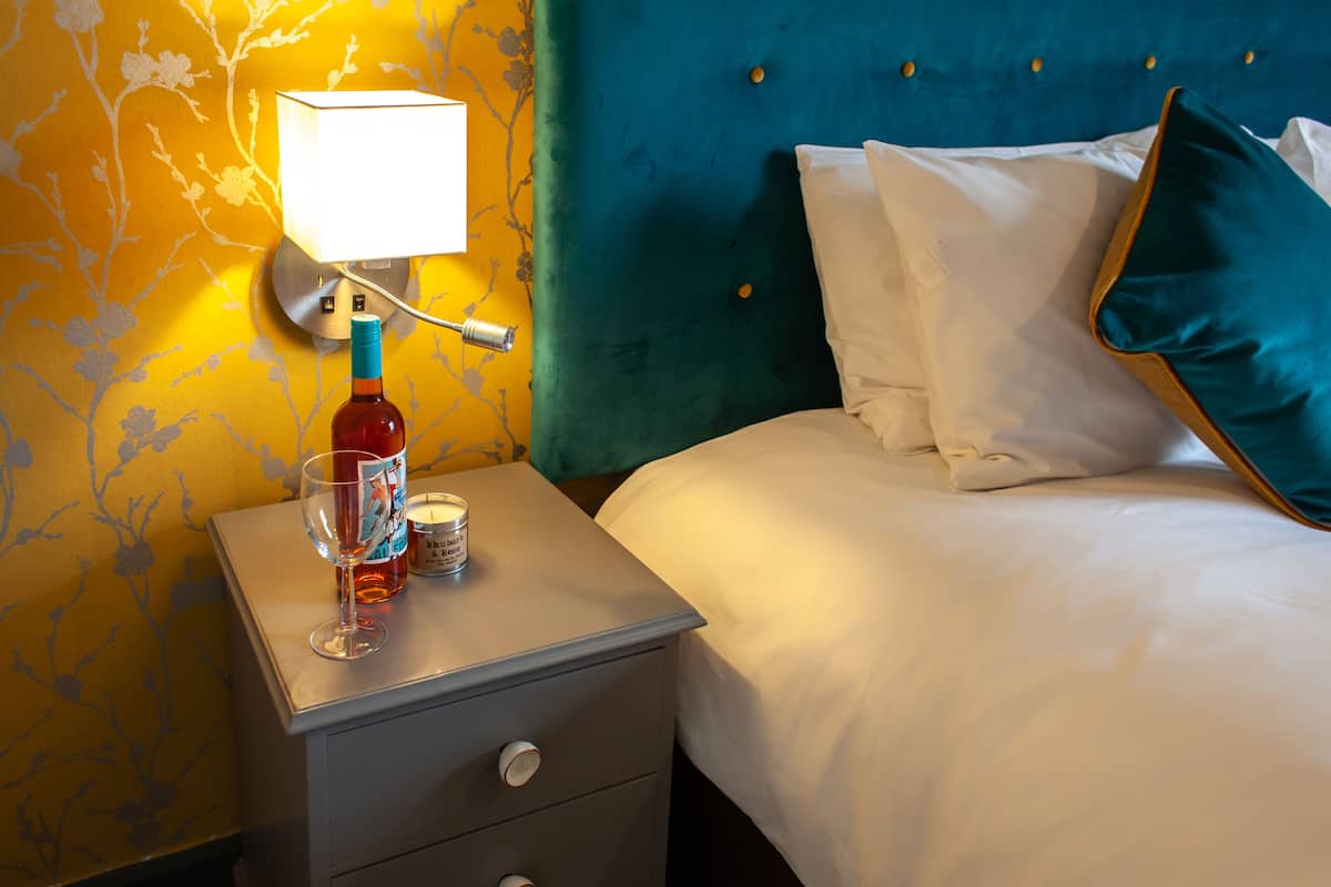 A neatly made bed with white linens and teal pillows, a bedside lamp, and a bottle of water on a nightstand.