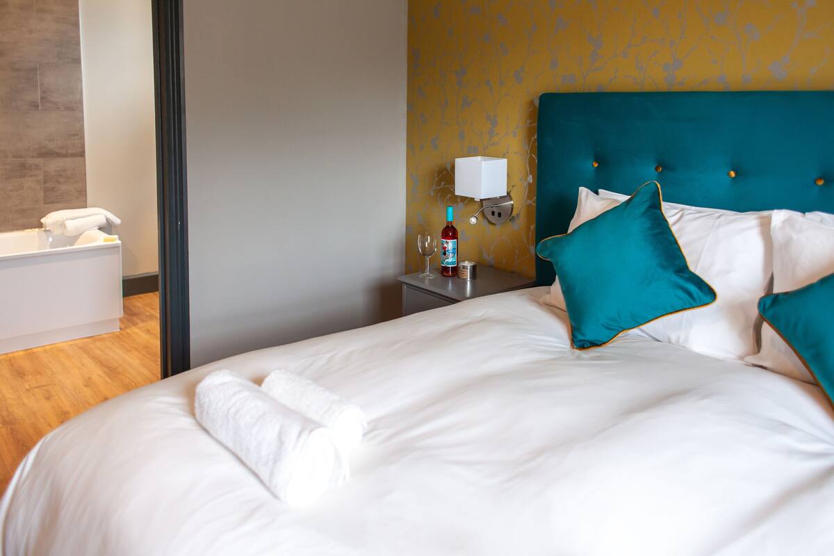 A neatly made bed with white linens and teal pillows, a bedside table with a bottle and glass, and a wall-mounted light fixture.