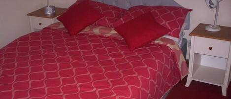 Una cama bien tendida con una colcha roja y beige estampada, dos almohadas rojas y un cabecero gris. Hay dos mesitas de noche con lámparas, una a cada lado de la cama.