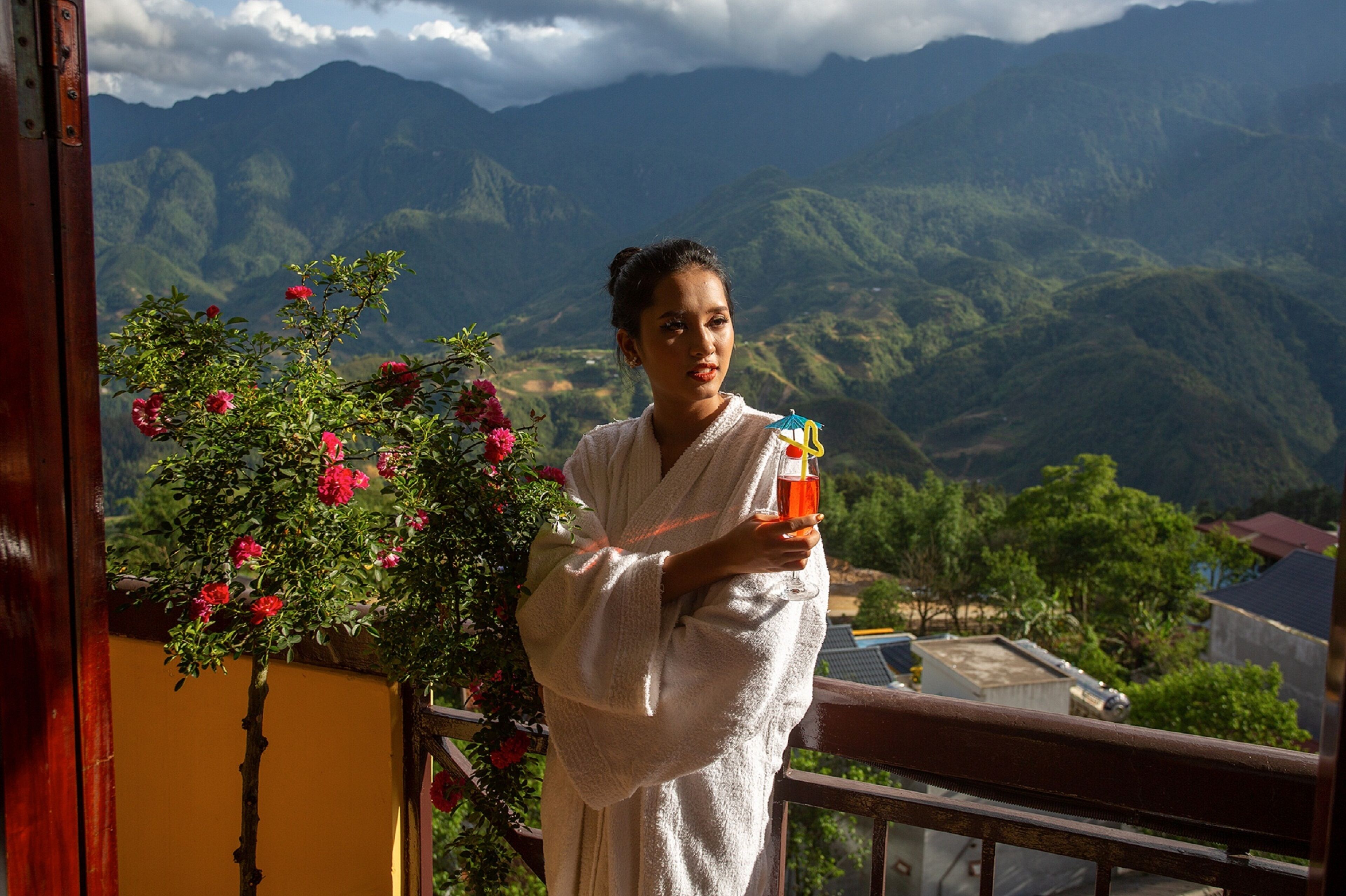Une personne vêtue d’une longue robe blanche, debout sur un balcon, d’où l’on peut admirer des montagnes et une végétation luxuriante.