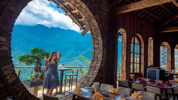 Un restaurant offrant une vue sur les montagnes, avec des arches en pierre et des poutres en bois. Les tables sont dressées avec des serviettes et un vase.