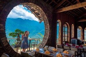 A restaurant with a view of mountains, stone arches, and wooden beams. Tables are set with napkins and a vase. - Chau Long Sapa 2 Hotel (Sa Pa)