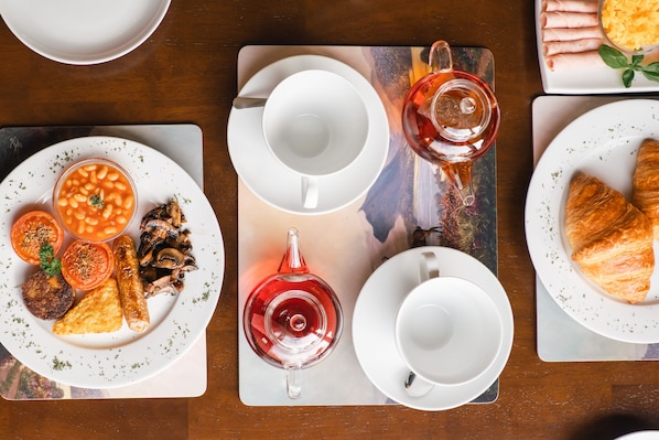 A breakfast setting with plates of beans, tomatoes, mushrooms, sausages, and croissants, along with cups and a teapot.