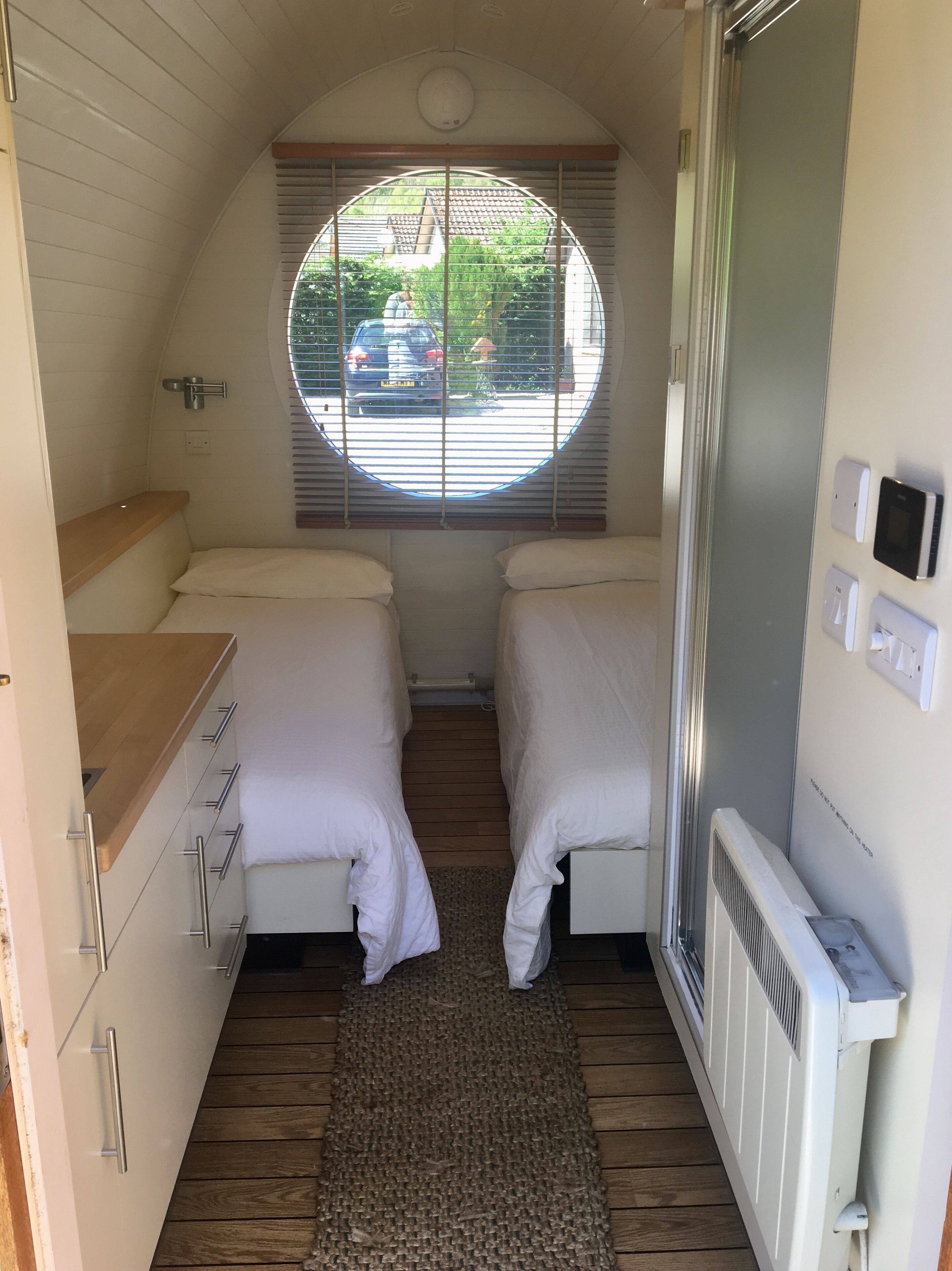 A compact bedroom with two beds, a window with blinds, a radiator, and a small kitchenette area.