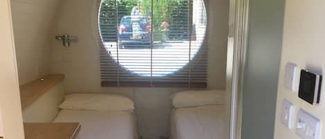 A compact bedroom with two beds, a window with blinds, a radiator, and a small kitchenette area.