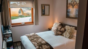 Un dormitorio con cama, ventana con vista a un edificio, cuadro enmarcado en la pared y mesita de noche con lámpara.