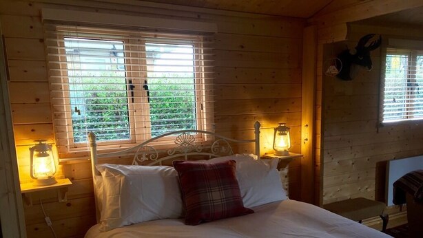 A wooden cabin bedroom with a bed, two bedside lamps, and a window with blinds. - Fourwinds Leisure (March)