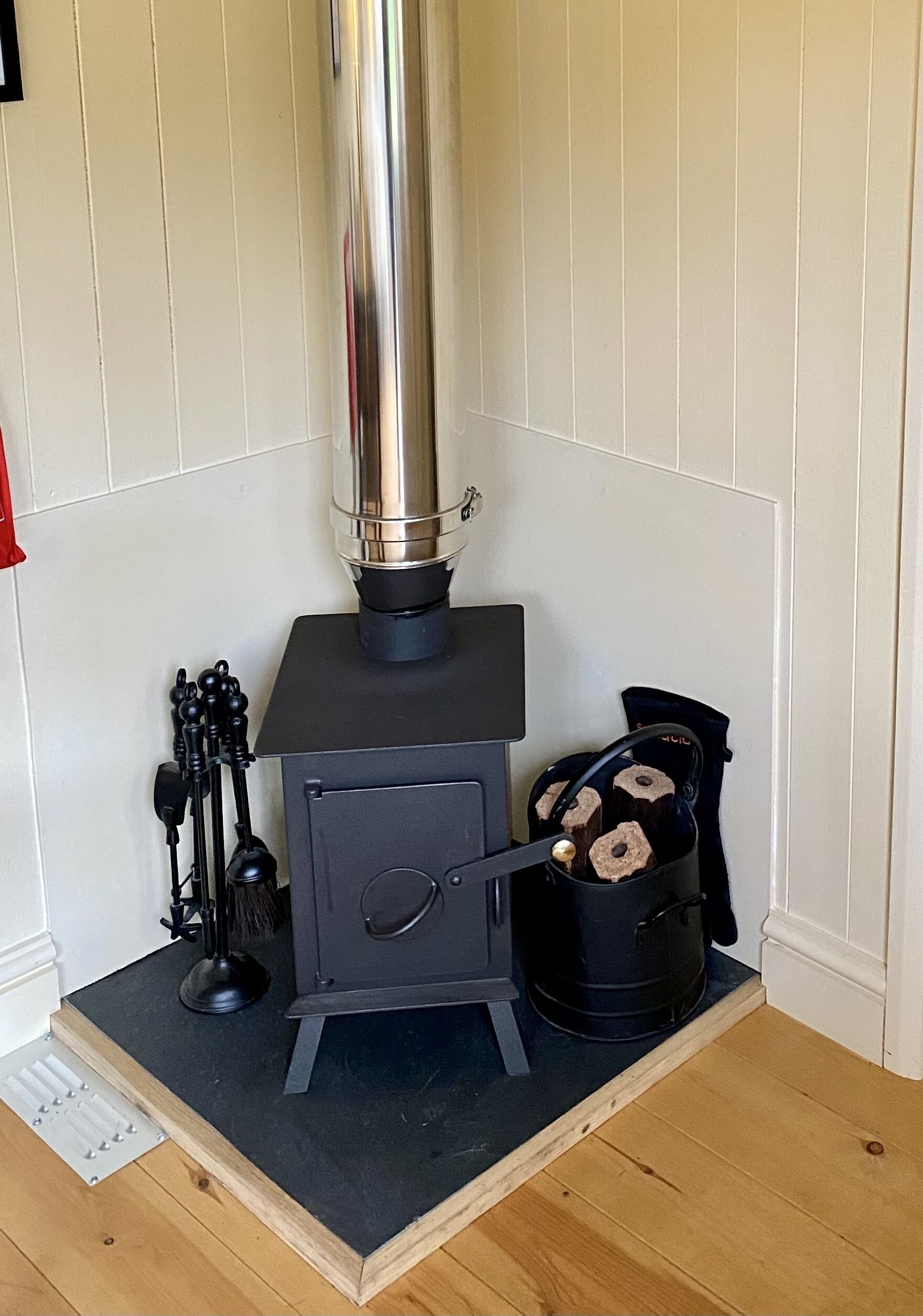A wood stove with a chimney, a pot with logs, and a brush on a wooden platform in a room with white walls.