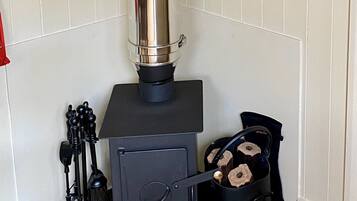 A wood stove with a chimney, a pot with logs, and a brush on a wooden platform in a room with white walls.
