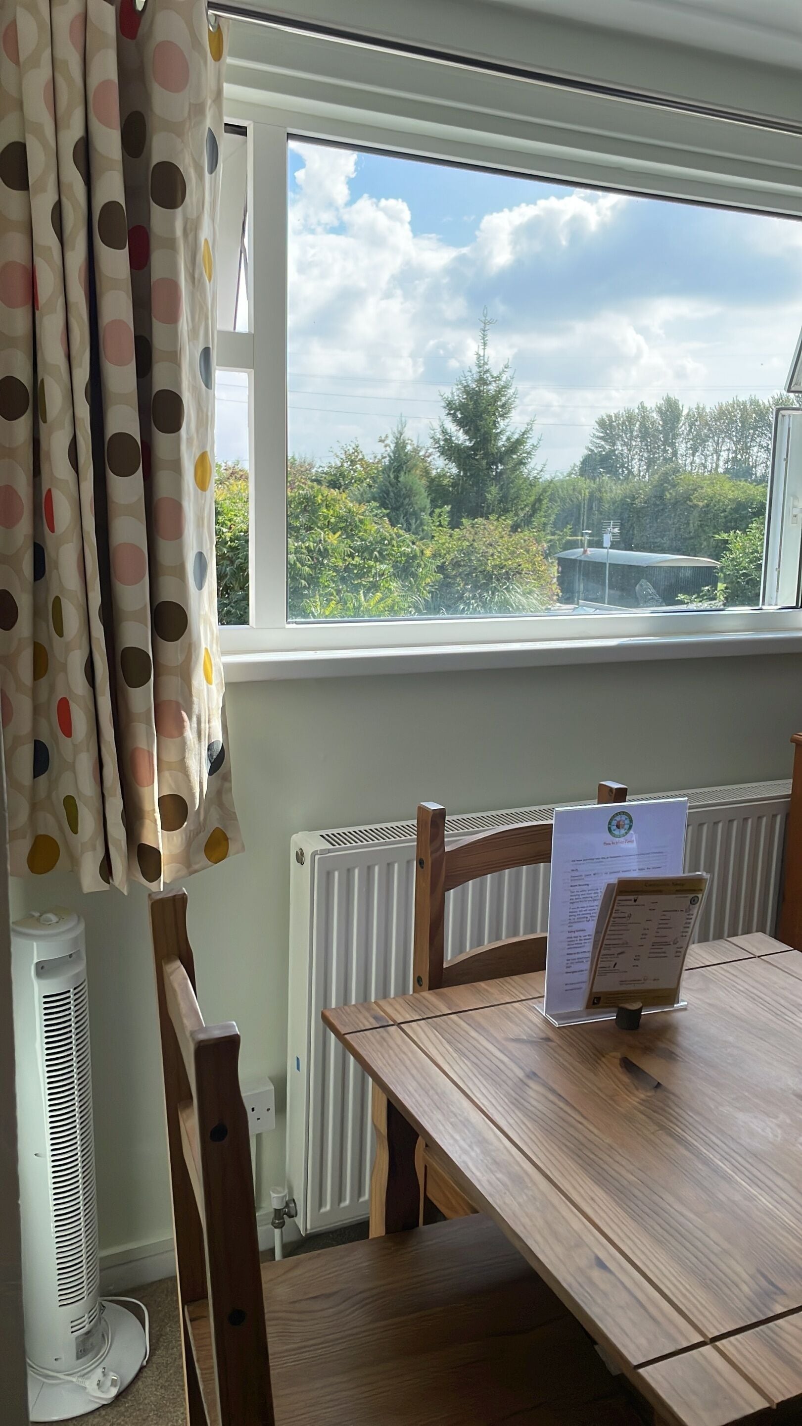 Un comedor con una mesa de madera, sillas, un atril para el menú, un radiador blanco y una ventana con vistas a zonas verdes y un árbol.
