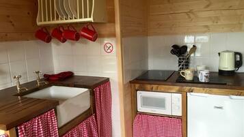 A compact kitchen with a sink, microwave, and refrigerator. There are red mugs hanging on the wall and a dish rack above the sink.