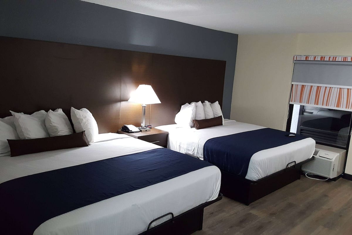 A hotel room with two beds, a bedside table with a lamp, a TV, and a window with blinds.