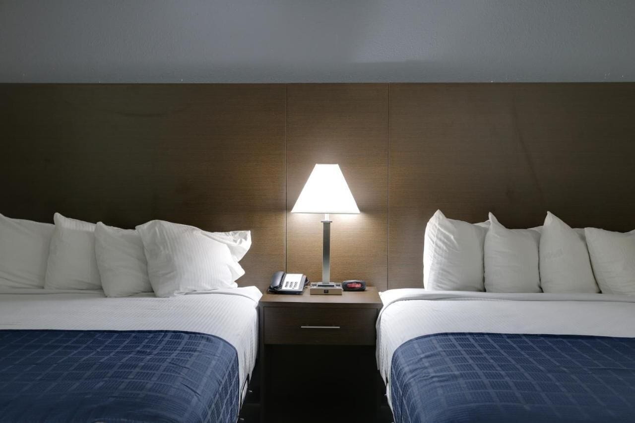 Una habitación de hotel con dos camas, una mesita de noche con lámpara, teléfono y reloj.