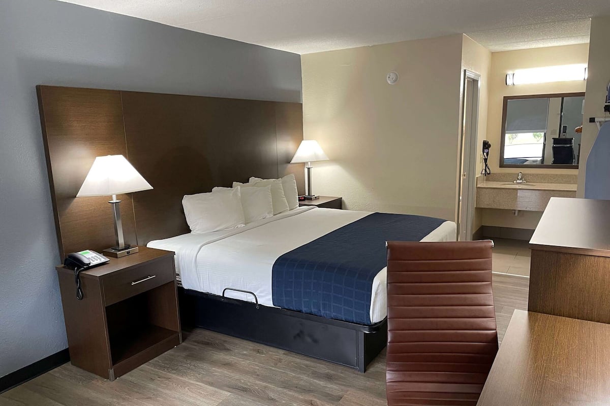 A hotel room with a bed, two bedside lamps, a nightstand with a phone, a desk with a chair, and a bathroom with a mirror and sink.