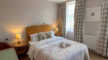 Quarto de hotel com cama, luminárias de cabeceira, uma cadeira, janela com cortinas e radiador.