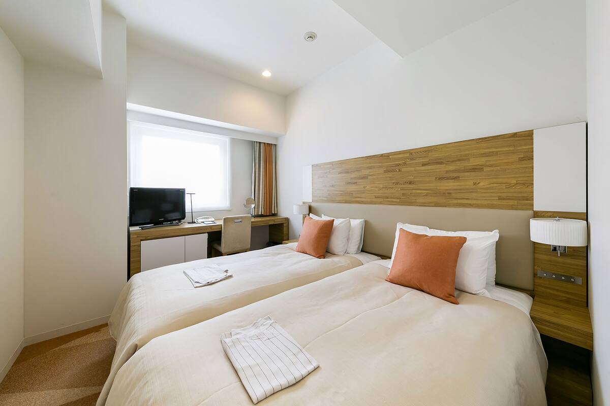 A hotel room with two beds, a desk with a TV, and a window with curtains.