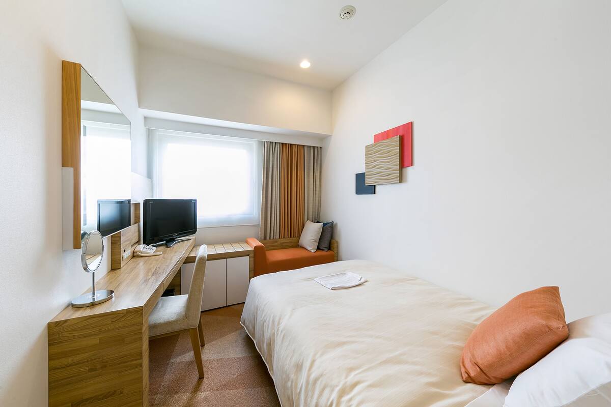 A hotel room with a bed, a desk with a computer, a chair, a mirror, and a window with curtains.