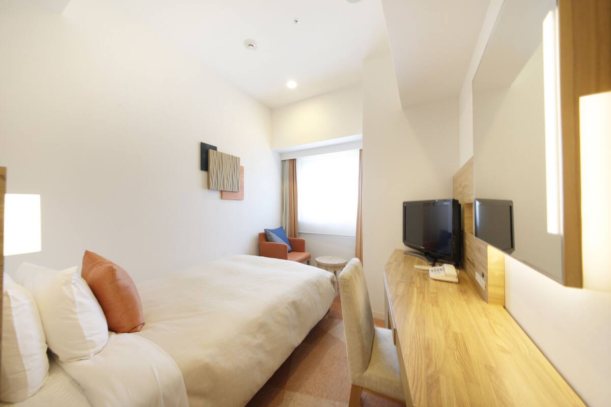 A hotel room with a bed, a desk with a TV, a chair, and a window.