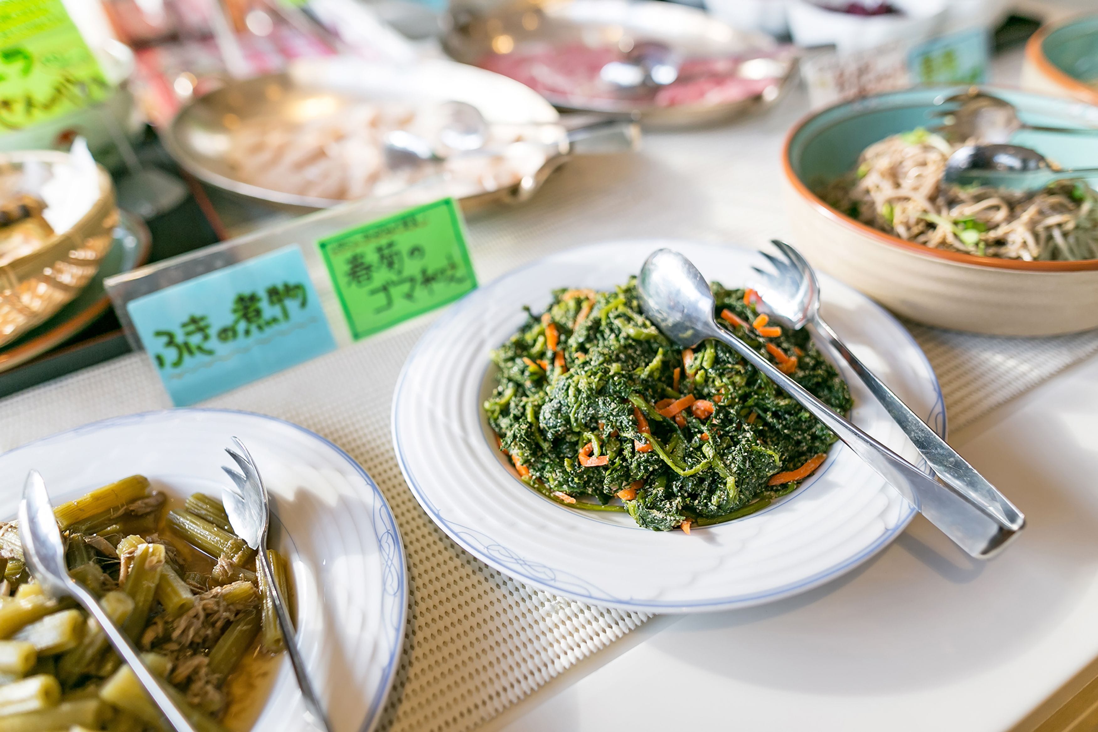 A table with various dishes, including a plate of greens and a bowl of noodles.